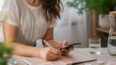 Frau schreibt an einem Schreibtisch in einen Kalender und hält dabei ein Smartphone